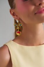 Model wearing Mirage Face Gold Earrings with abstract face design, blue eyes and red lips