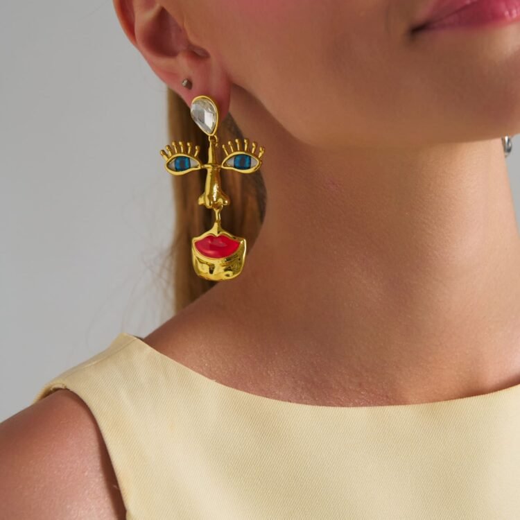 Model wearing Mirage Face Gold Earrings with abstract face design, blue eyes and red lips